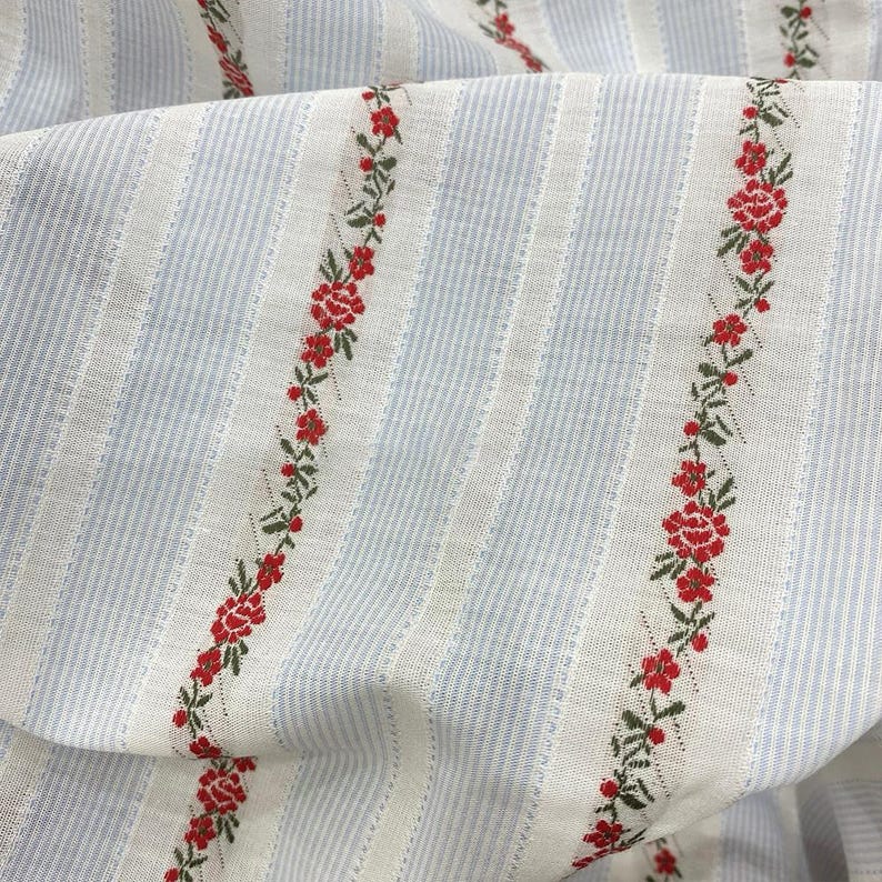 May include: Close-up of a fabric with a striped pattern in light blue and white. Vertical rows of red floral embroidery with green leaves run along the fabric. The fabric is folded, showing its texture and design.