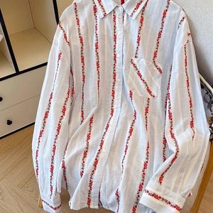 May include: A light blue and white striped button-down shirt with long sleeves. The shirt features vertical lines of red floral embroidery. The collar, pocket, and cuffs are also adorned with the same red floral pattern.