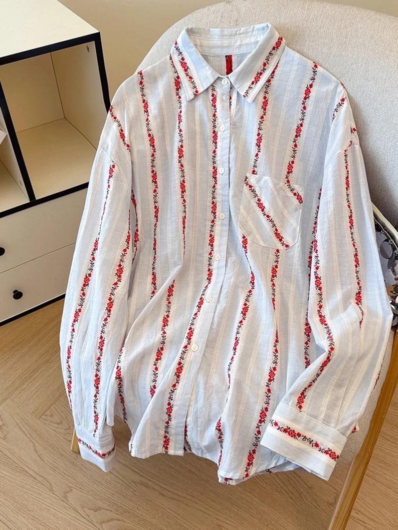 May include: A light blue and white striped button-down shirt with long sleeves. The shirt features vertical lines of red floral embroidery. The collar, pocket, and cuffs are also adorned with the same red floral pattern.