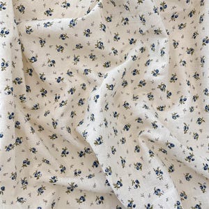 May include: Off-white fabric featuring a small floral design. The pattern consists of tiny blue flowers and green leaves on a cream background. The fabric appears to be textured, possibly linen or a similar natural fibre.