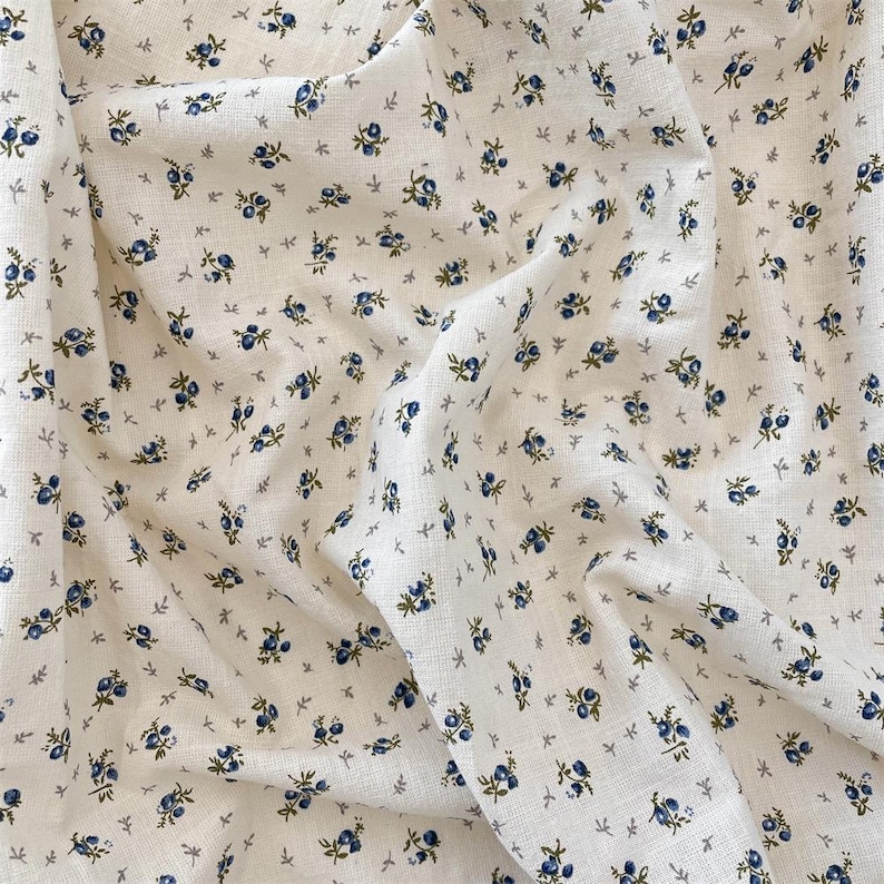 May include: Off-white fabric featuring a small floral design. The pattern consists of tiny blue flowers and green leaves on a cream background. The fabric appears to be textured, possibly linen or a similar natural fibre.