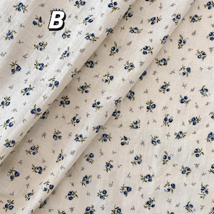May include: Cream-coloured fabric featuring a repeating pattern of small blue floral clusters and tiny grey leaf shapes. The letter "B" is printed in black and white in the upper left corner. The fabric is folded, showing its texture.