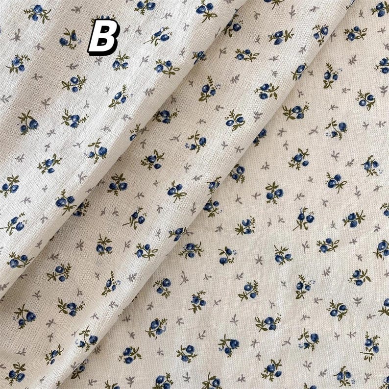 May include: Cream-coloured fabric featuring a repeating pattern of small blue floral clusters and tiny grey leaf shapes. The letter "B" is printed in black and white in the upper left corner. The fabric is folded, showing its texture.