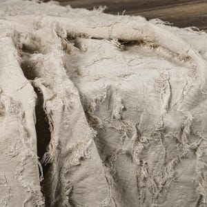 May include: A beige fabric throw blanket featuring a textured surface with frayed, raw-edge detailing arranged in a repeating pattern across the material. The textile has a soft, wrinkled appearance with loose threads throughout the design.