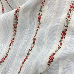 May include: Close-up of a fabric with a striped pattern in white and light blue. Red floral embroidery with green leaves runs vertically along the fabric. The fabric is slightly wrinkled, suggesting it is a soft material.
