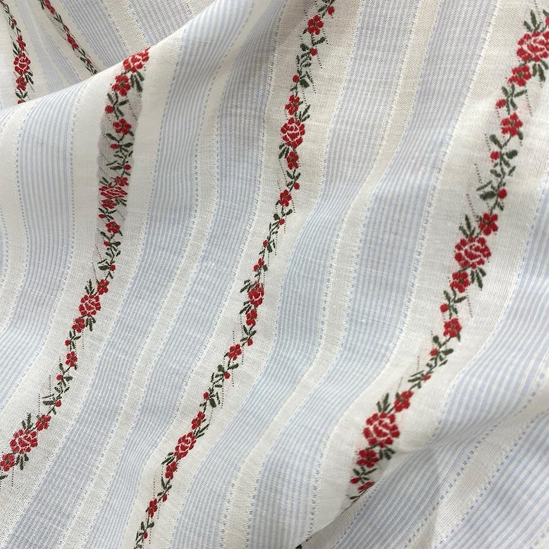 May include: Close-up of a fabric with a striped pattern in white and light blue. Red floral embroidery with green leaves runs vertically along the fabric. The fabric is slightly wrinkled, suggesting it is a soft material.