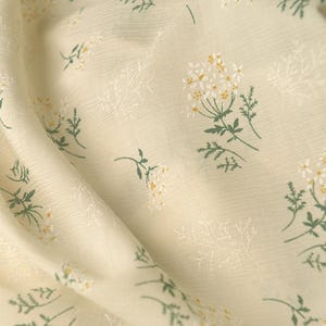 Op de afbeelding: Een lichtcr&egrave;mekleurige stof met een herhalend patroon van kleine witte en gele bloemen met groene stengels en bladeren. Het textiel heeft een subtiele, fijn geribbelde textuur over het geheel, met het bloemmotief op het oppervlak gedrukt.