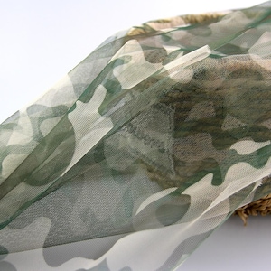 May include: A sheer, lightweight fabric featuring a camouflage pattern in shades of olive green, dark green, and off-white. The material has a fine mesh texture and is draped over a woven, textured brown object, showing semi-transparency.