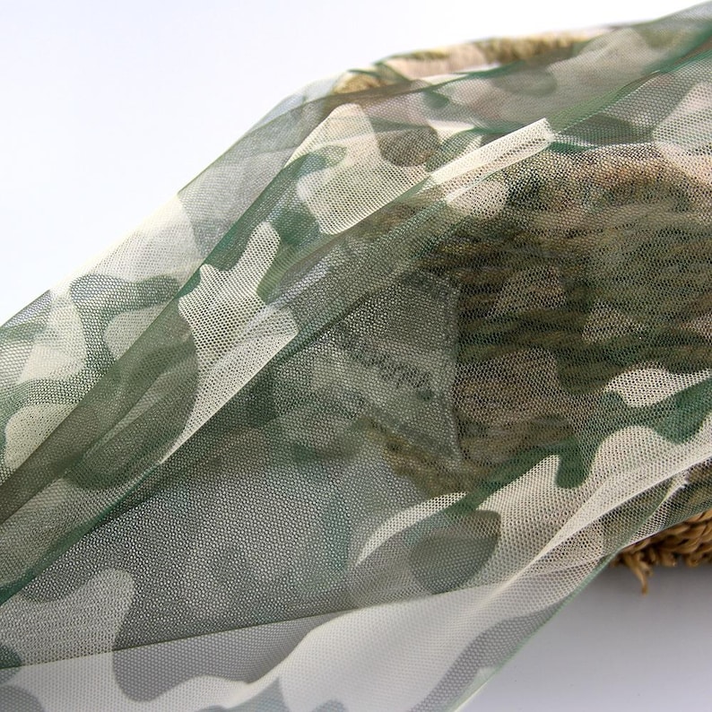 May include: A sheer, lightweight fabric featuring a camouflage pattern in shades of olive green, dark green, and off-white. The material has a fine mesh texture and is draped over a woven, textured brown object, showing semi-transparency.