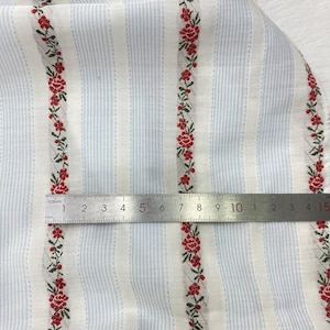 May include: Close-up of fabric with a light blue and white striped pattern. Vertical rows of red floral designs with green leaves are interspersed between the stripes. A metal ruler is placed across the fabric.
