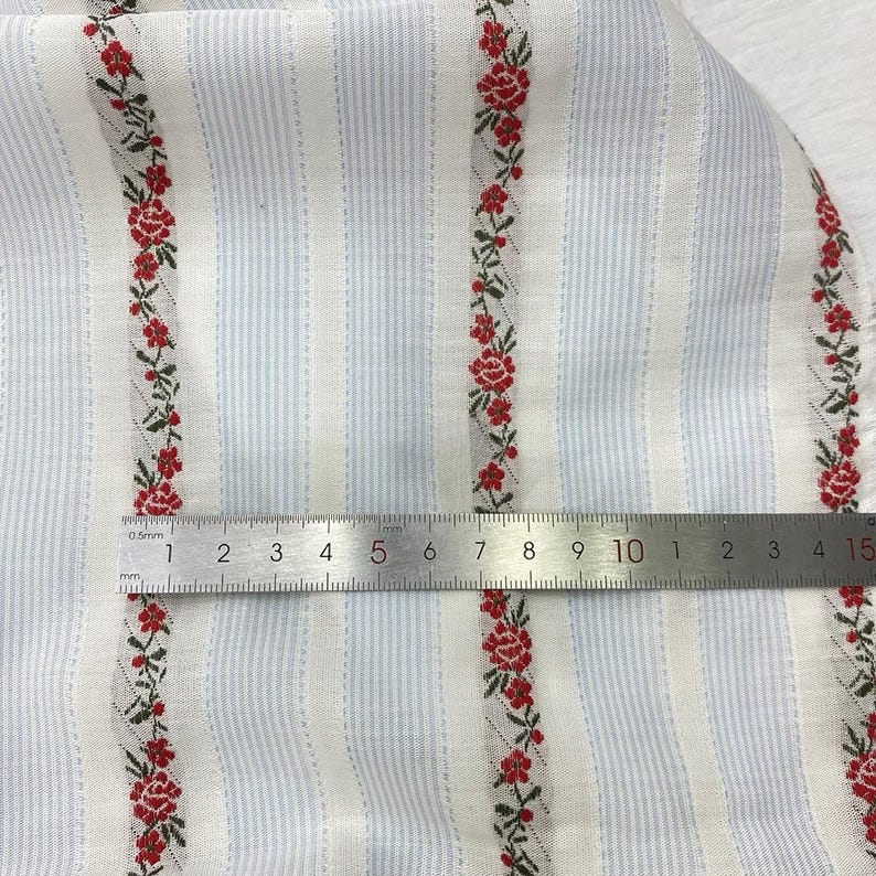 May include: Close-up of fabric with a light blue and white striped pattern. Vertical rows of red floral designs with green leaves are interspersed between the stripes. A metal ruler is placed across the fabric.