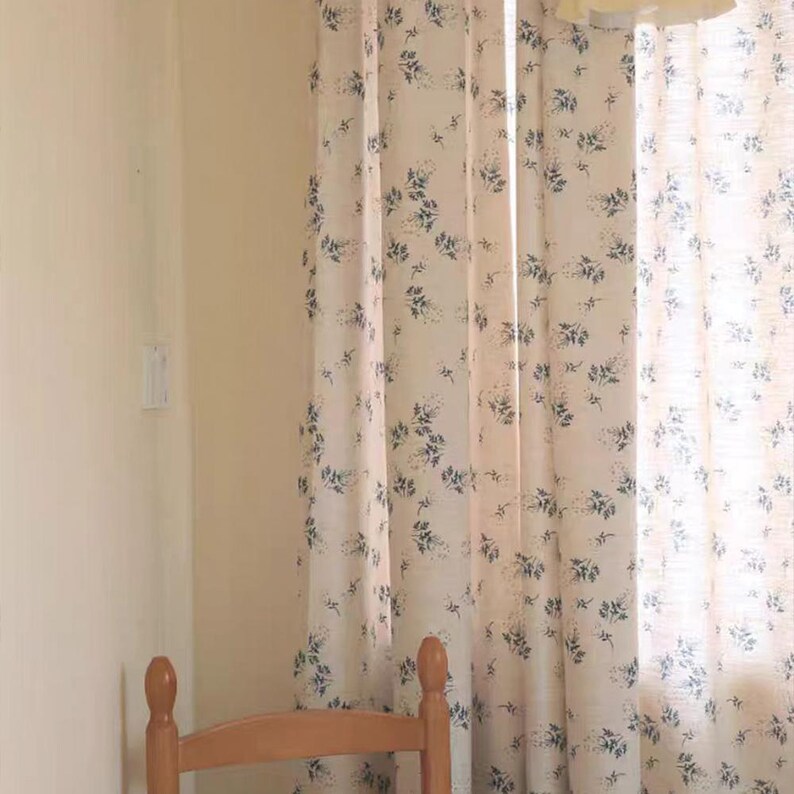 Op de afbeelding: Vloerlange gordijnen van licht cr&egrave;mekleurige stof met een herhalend patroon van kleine, donkerblauwe bloemtakjes en blaadjes. De gordijnen hangen in verticale plooien voor een raam, waarbij de rugleuning van een houten stoel zichtbaar is.