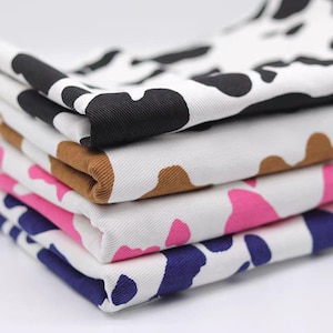 Può includere: Una pila di pezzi di tessuto piegati con un motivo a macchie di mucca. Lo strato superiore presenta macchie nere su bianco, il secondo macchie marroni su bianco, il terzo macchie rosa su bianco e la base macchie blu scuro su fondo bianco.