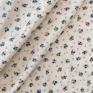 May include: Cream-coloured fabric featuring a repeating pattern of small blue floral clusters and tiny grey leaves. The fabric is folded, displaying the texture and design. The overall aesthetic is delicate and vintage-inspired.