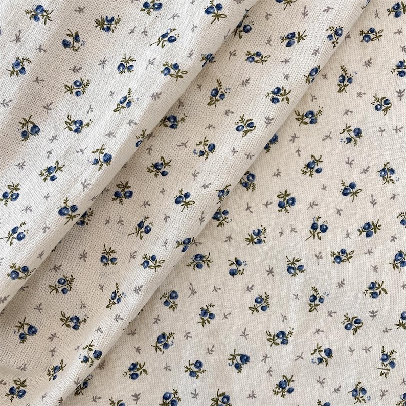 May include: Cream-coloured fabric featuring a repeating pattern of small blue floral clusters and tiny grey leaves. The fabric is folded, displaying the texture and design. The overall aesthetic is delicate and vintage-inspired.