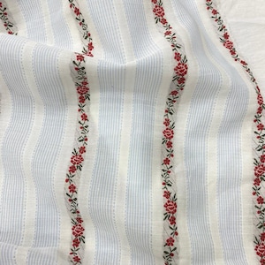 May include: Close-up of a fabric with vertical stripes in light blue and white. Red floral embroidery with green leaves runs vertically along the fabric. The fabric has a soft, textured appearance.