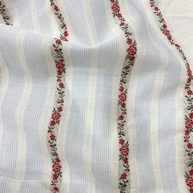 May include: Close-up of a fabric with vertical stripes in light blue and white. Red floral embroidery with green leaves runs vertically along the fabric. The fabric has a soft, textured appearance.