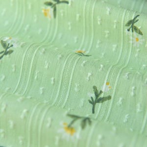 Op de afbeelding: Een lichtgroene stof met een herhalend patroon van kleine witte madeliefjes met gele harten en groene stelen. Het materiaal heeft een getextureerd oppervlak met verticale strepen en kleine verhoogde stippen, wat zorgt voor een subtiel, tactiel effect.