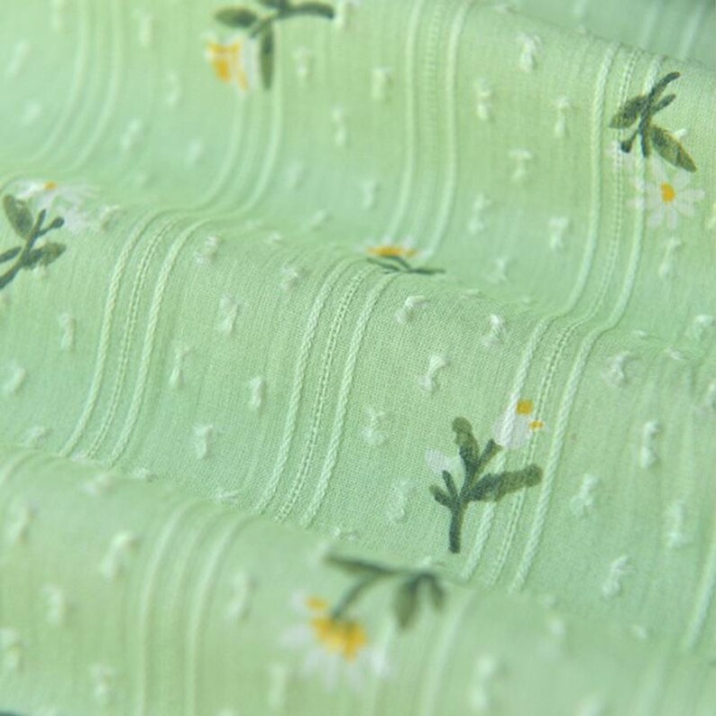 Op de afbeelding: Een lichtgroene stof met een herhalend patroon van kleine witte madeliefjes met gele harten en groene stelen. Het materiaal heeft een getextureerd oppervlak met verticale strepen en kleine verhoogde stippen, wat zorgt voor een subtiel, tactiel effect.