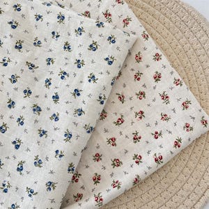 May include: Two pieces of off-white fabric with floral patterns. One fabric features blue berry-like flowers, while the other has red and pink rose-like flowers. Both fabrics have small grey cross-shaped accents.