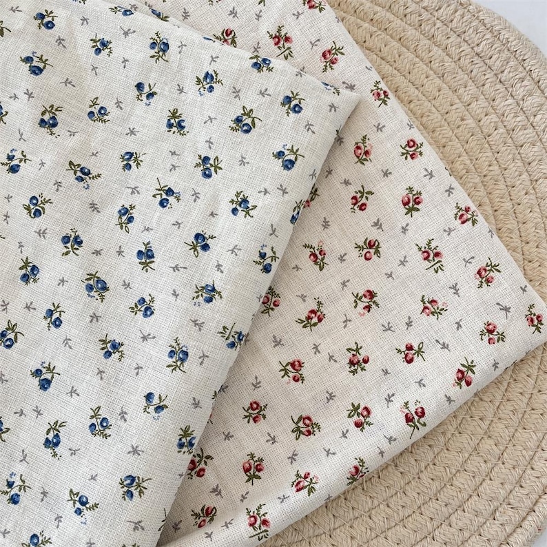 May include: Two pieces of off-white fabric with floral patterns. One fabric features blue berry-like flowers, while the other has red and pink rose-like flowers. Both fabrics have small grey cross-shaped accents.