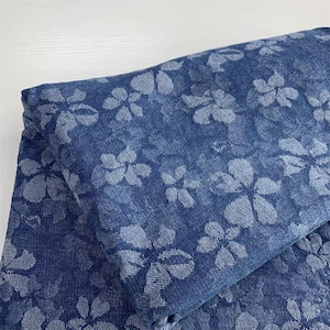 Puede incluir: Tela vaquera doblada con un estampado floral. La tela es de color azul oscuro con un diseño floral azul más claro. Las flores están espaciadas uniformemente por toda la tela.