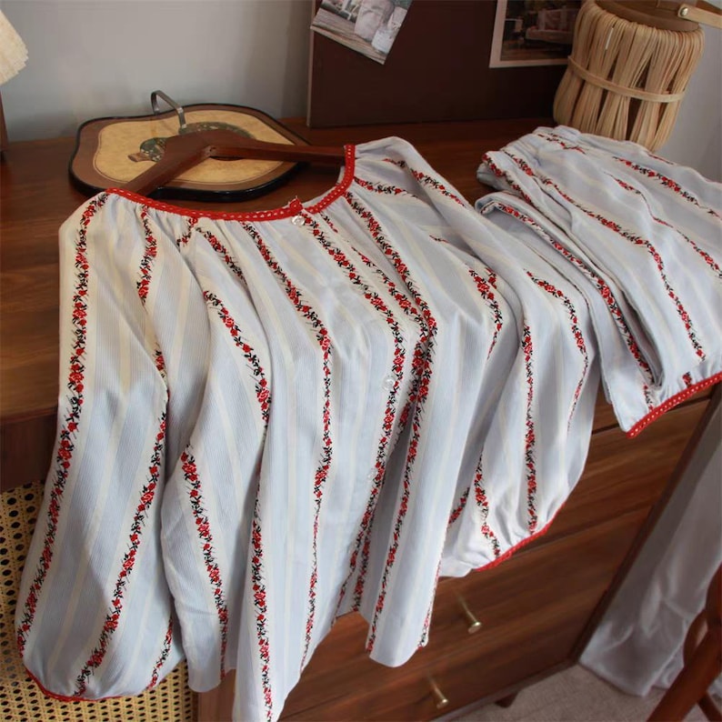 May include: A light blue pajama set with vertical stripes and red floral embroidery. The long-sleeved top and matching pants have red trim. The set is displayed on a wooden dresser.