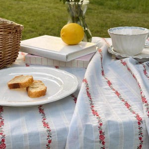 May include: A picnic scene with a white tablecloth featuring red floral embroidery. A white plate holds two slices of bread. A lemon sits atop a stack of books. A teacup and a wicker basket are also visible.
