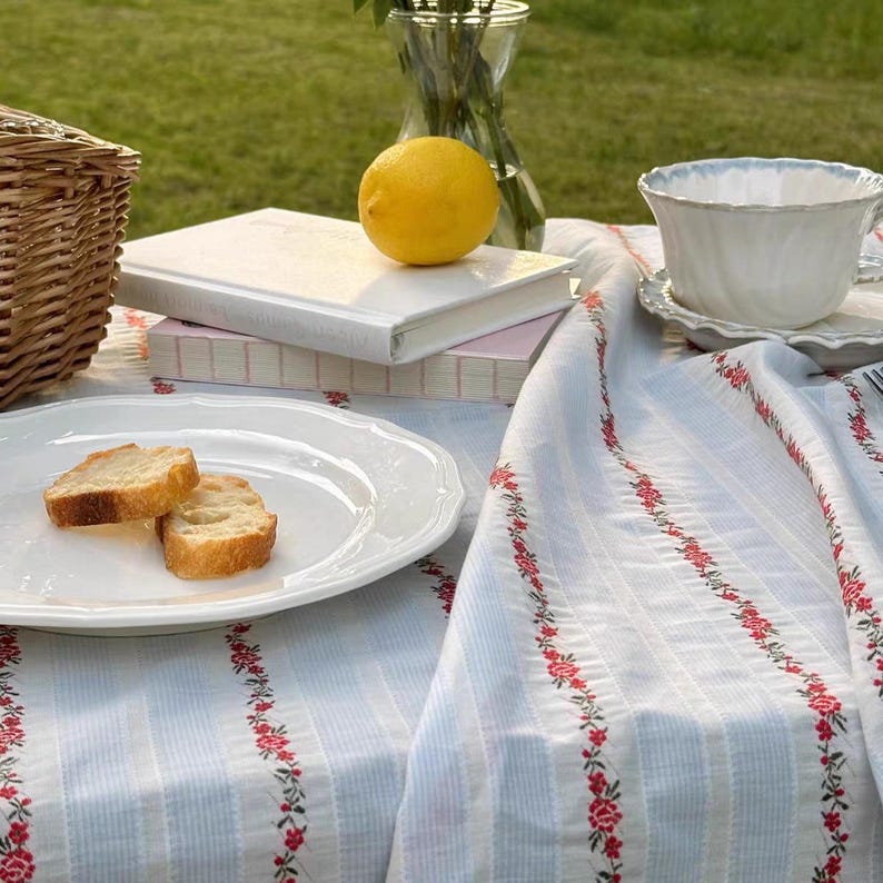 May include: A picnic scene with a white tablecloth featuring red floral embroidery. A white plate holds two slices of bread. A lemon sits atop a stack of books. A teacup and a wicker basket are also visible.