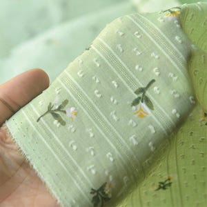 Op de afbeelding: Een lichtgroene stof met een delicaat bloemenpatroon van kleine witte en gele bloemetjes, geaccentueerd door verticale strepen en een gestructureerd stippeleffect. Het materiaal is semi-transparant en lichtgewicht met een zachte weving.