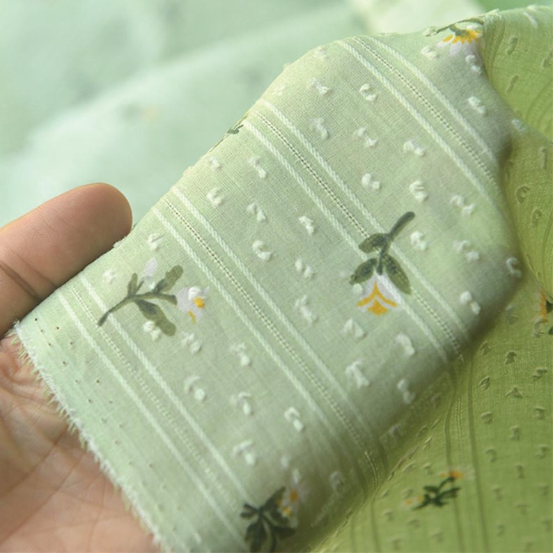 Op de afbeelding: Een lichtgroene stof met een delicaat bloemenpatroon van kleine witte en gele bloemetjes, geaccentueerd door verticale strepen en een gestructureerd stippeleffect. Het materiaal is semi-transparant en lichtgewicht met een zachte weving.