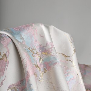 Puede incluir: Una pieza de tela con una base lisa de color blanco roto y un patr&oacute;n de m&aacute;rmol abstracto en tonos pastel de rosa y azul claro. El dise&ntilde;o est&aacute; acentuado con detalles de l&aacute;mina de oro met&aacute;lico que siguen las formas fluidas y org&aacute;nicas.