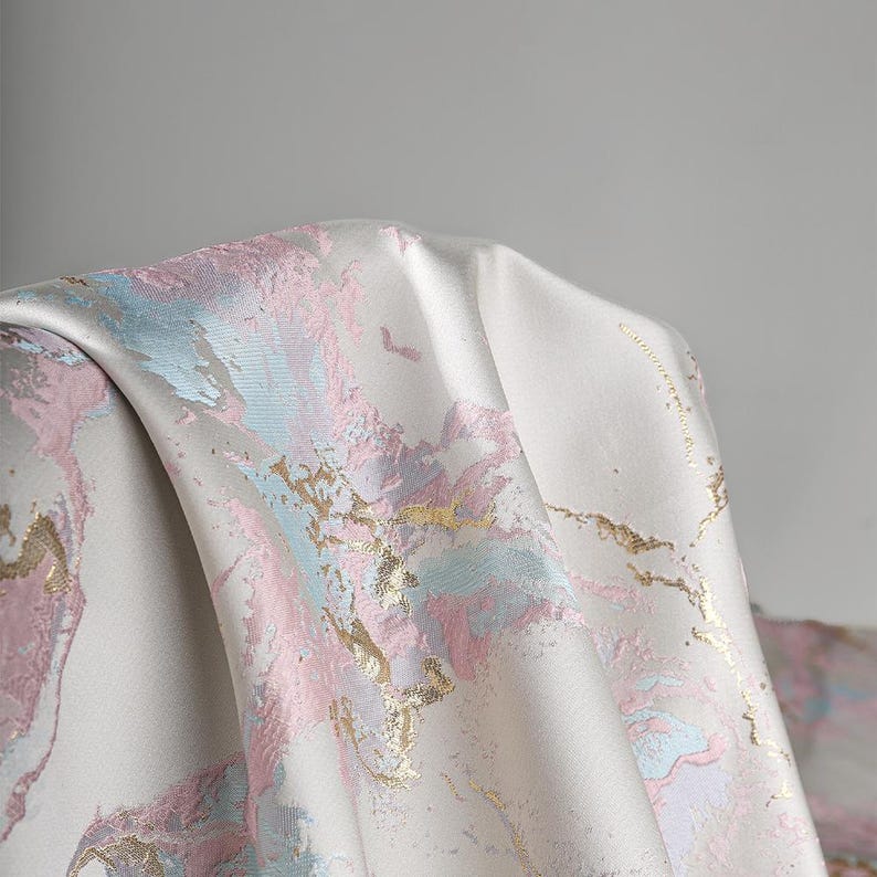 Puede incluir: Una pieza de tela con una base lisa de color blanco roto y un patr&oacute;n de m&aacute;rmol abstracto en tonos pastel de rosa y azul claro. El dise&ntilde;o est&aacute; acentuado con detalles de l&aacute;mina de oro met&aacute;lico que siguen las formas fluidas y org&aacute;nicas.