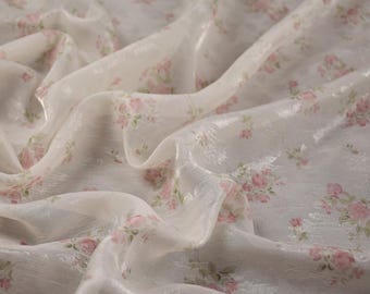 Puede incluir: Una tela transparente de color blanco roto con un patrón repetitivo de pequeños grupos de rosas de color rosa suave con hojas verdes apagadas. El material tiene un acabado texturizado y sutilmente brillante que capta la luz y crea pliegues.