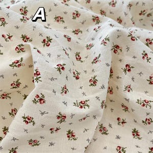 May include: Cream-coloured fabric featuring a floral design. Small clusters of red and green flowers are spread across the material, interspersed with tiny grey leaf and star-shaped details. The letter "A" is visible in the upper left corner.