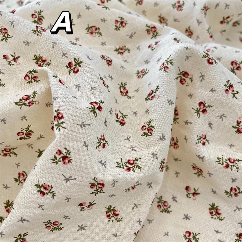 May include: Cream-coloured fabric featuring a floral design. Small clusters of red and green flowers are spread across the material, interspersed with tiny grey leaf and star-shaped details. The letter "A" is visible in the upper left corner.