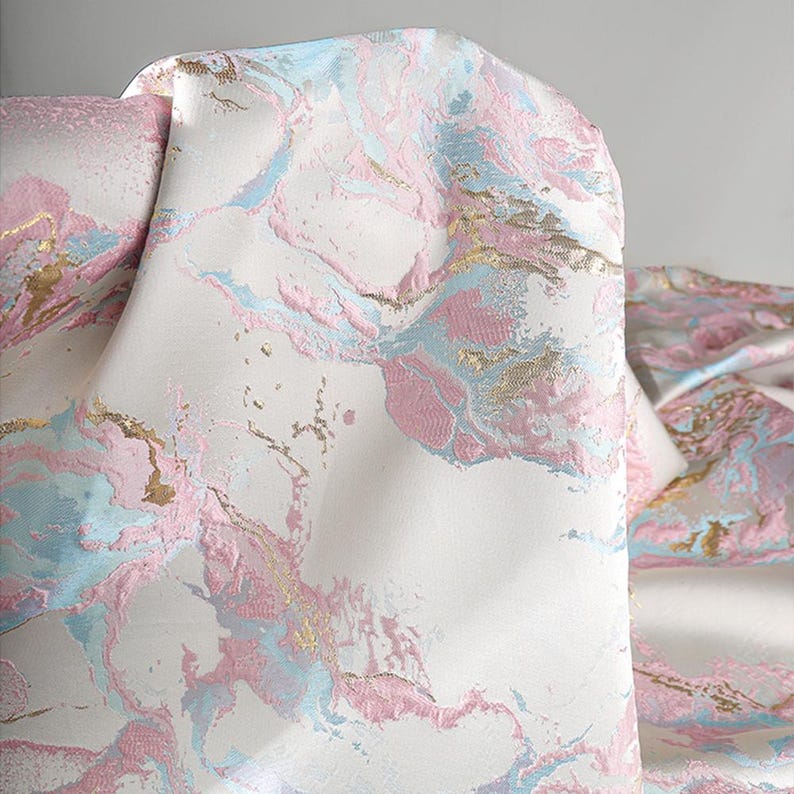 Puede incluir: Una tela con un patr&oacute;n de m&aacute;rmol sobre una base blanca, con remolinos de color rosa suave y azul claro, y acentos de oro met&aacute;lico. El material tiene una superficie texturizada con &aacute;reas elevadas que crean profundidad y dimensi&oacute;n en el tejido.