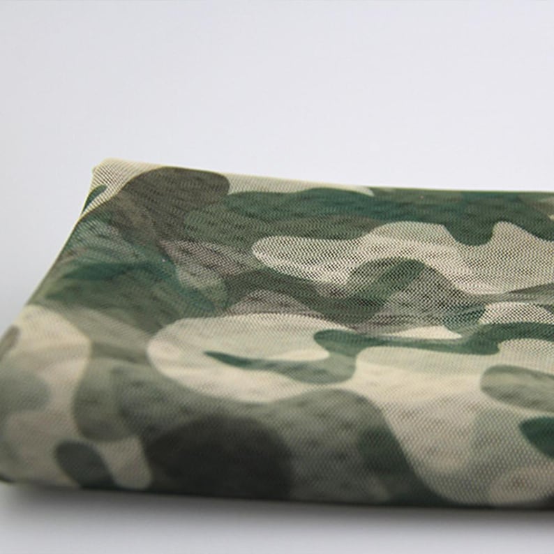 May include: A piece of semi-sheer mesh fabric featuring a camouflage pattern in shades of olive green, dark forest green, and light beige. The material has a fine, grid-like texture visible across the surface, creating a lightweight appearance.