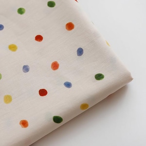 May include: A folded piece of off-white fabric featuring a scattered pattern of small, hand-drawn style polka dots in various colors including red, orange, yellow, green, and blue. The textile has a smooth, woven texture with visible fibers.