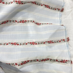 May include: White and light blue striped fabric with red floral embroidery. The fabric has a textured appearance and a fringed edge. The floral embroidery is a repeating pattern of red flowers and green leaves.