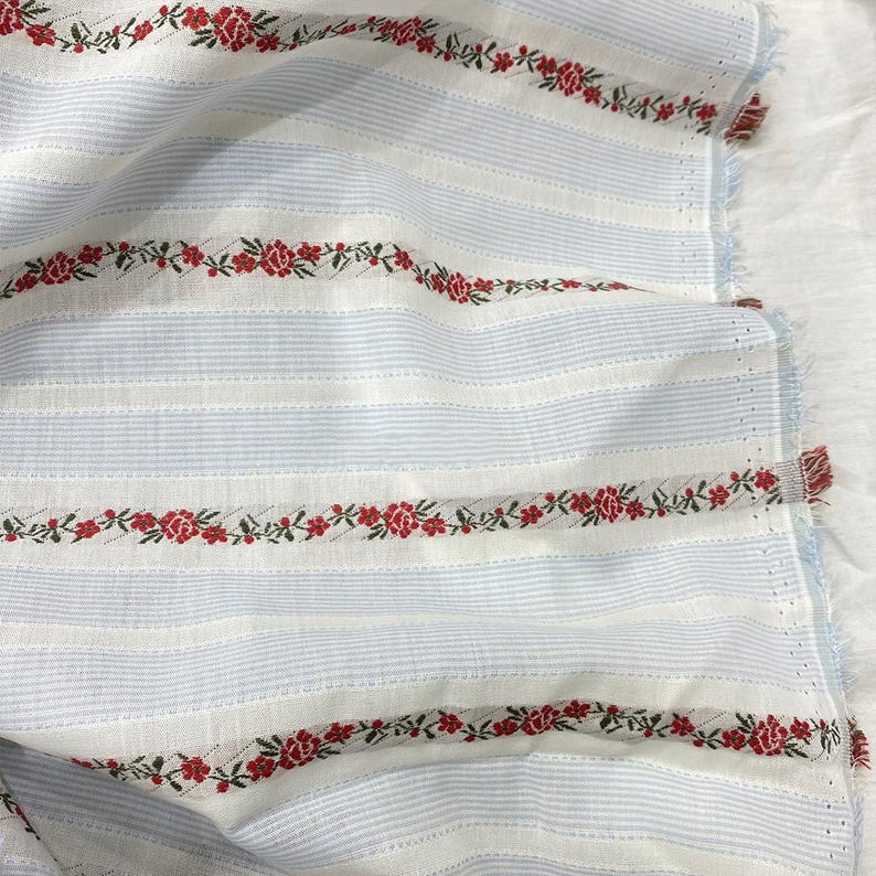 May include: White and light blue striped fabric with red floral embroidery. The fabric has a textured appearance and a fringed edge. The floral embroidery is a repeating pattern of red flowers and green leaves.