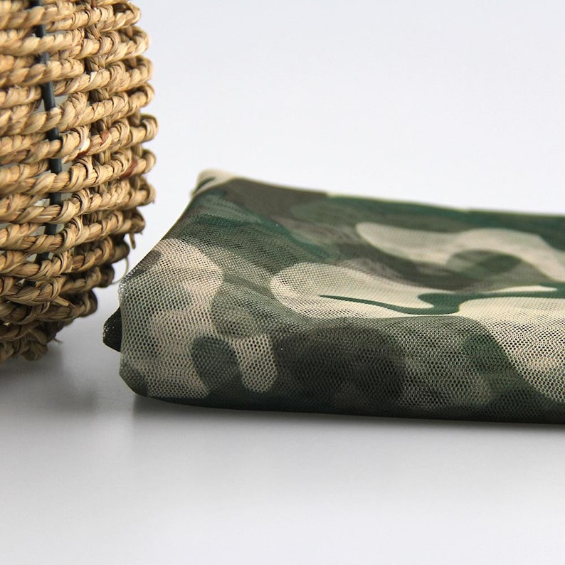 May include: A folded piece of sheer mesh fabric featuring a camouflage pattern in shades of dark green, olive, and light beige. The material has a fine, grid-like texture and is layered, showing the semi-transparent quality of the textile.