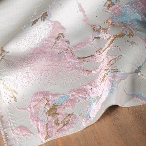 Puede incluir: Una pieza de tela con base blanca y un patr&oacute;n abstracto en hilos de color rosa suave, azul claro y oro met&aacute;lico. El material tiene una apariencia tejida y texturizada con un brillo sutil, mostrando formas org&aacute;nicas y l&iacute;neas fluidas.