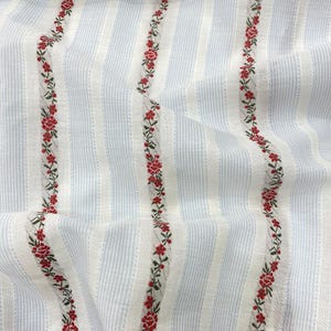 May include: Close-up of a fabric with a striped pattern in light blue and white. Vertical rows of red floral embroidery with green leaves run down the fabric, creating a delicate and textured design. The fabric has a soft, slightly wrinkled appearance.
