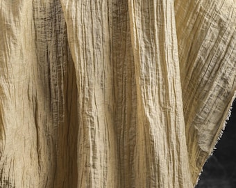Può includere: Un tessuto beige chiaro con una distinta consistenza stropicciata verticale. Il materiale presenta una serie di pieghe parallele in rilievo che corrono dall'alto verso il basso, creando un aspetto ondulato con bordi sfilacciati visibili sul lato.