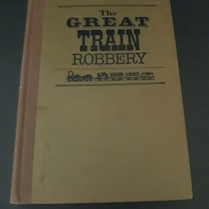 May include: A tan hardback book with the title "The Great Train Robbery" printed in black letters. A small black and white illustration of a train is below the title.