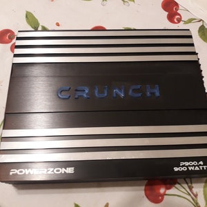 May include: A black car amplifier with silver accents. The amplifier has the text "CRUNCH" in blue letters and "POWERZONE" in white letters. The amplifier also has the text "P900.4 900 WATTS" in white letters.