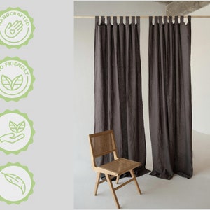 May include: Two dark grey linen curtains hanging from a wooden rod. The curtains have a simple design with a loop at the top of each panel. A wooden chair with a woven seat is in the foreground.