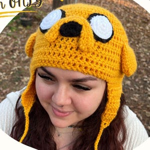 May include: A yellow crocheted hat featuring the character Jake the Dog with large white eyes, black pupils, a black nose, and two side flaps with dangling ties. The text Jake the Dog Hat Pattern Only and Crazy 8 Creations @Crazy8Creationss appear.