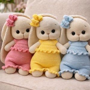 Puede incluir: Tres muñecas de conejito hechas a mano con ganchillo. Cada conejito lleva un atuendo de diferente color: rosa, amarillo y azul. Los conejitos tienen orejas grandes y caídas y lazos en la cabeza. Las muñecas están sentadas sobre una superficie beige.
